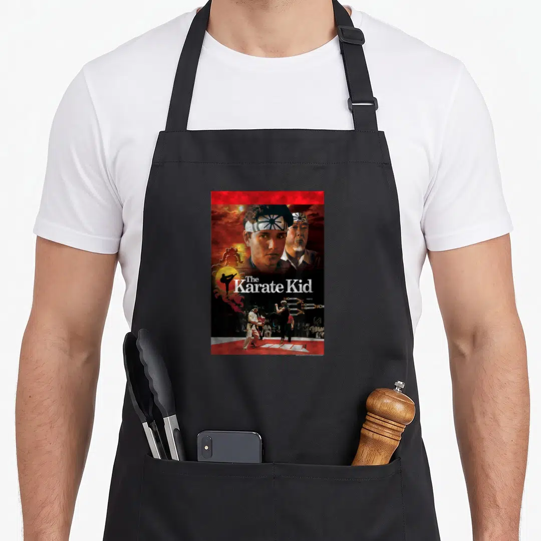 Karate distressed apron - The Karate Kid text over a sun with Daniel LaRusso and Mr. Miyagi portraits above a fight — shown on a black cotton canvas apron against a white background