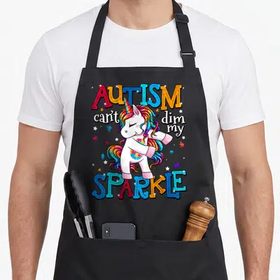 Autism awareness unicorn apron - Cartoon unicorn with rainbow mane surrounded by Autism Can't Dim My Sparkle text — shown on a black cotton canvas apron against a white background