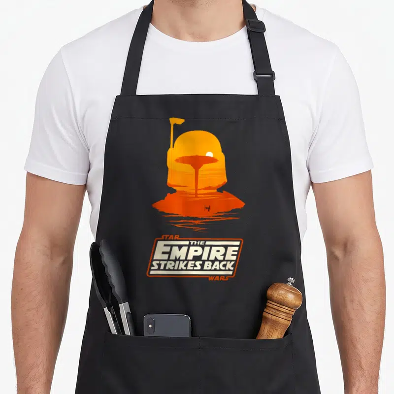 Empire boba apron - Orange helmet silhouette containing a landscape scene above Star Wars Empire Strikes Back text logo — shown on a black cotton canvas apron against a white background
