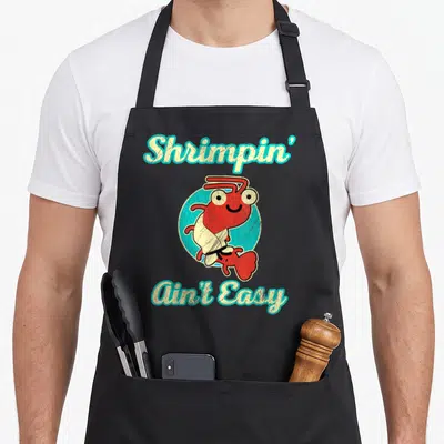 Jiu jitsu apron - Cartoon red shrimp in white gi inside blue circle with arched text Shrimpin Ain Easy — shown on a black cotton canvas apron against a white background