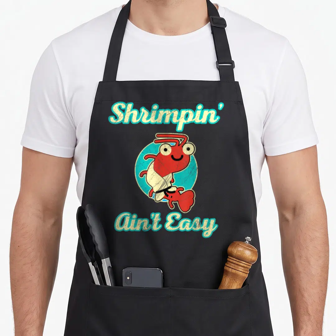 Jiu jitsu apron - Cartoon red shrimp in white gi inside blue circle with arched text Shrimpin Ain Easy — shown on a black cotton canvas apron against a white background