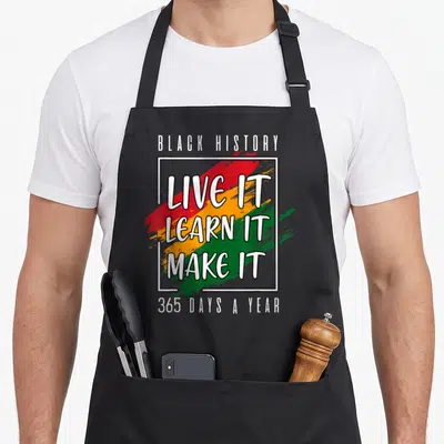 History novelty apron - Stacked text LIVE IT LEARN IT MAKE IT over red yellow green brush strokes with white square — shown on a black cotton canvas apron against a white background