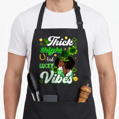 Thick st patrick day aprons - Cartoon woman in green hat surrounded by Thick thighs lucky vibes text, shamrocks — shown on a black cotton canvas apron against a white background