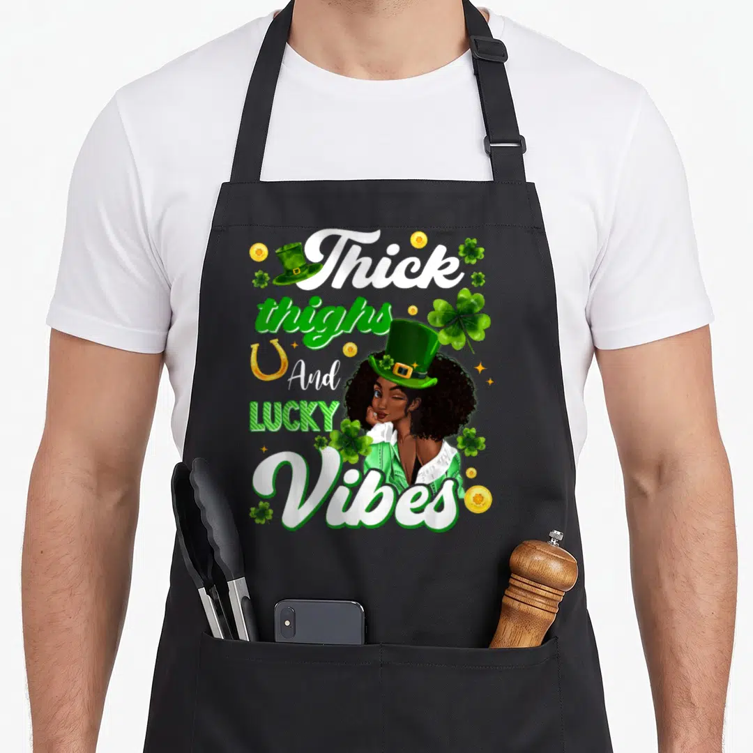 Thick st patrick day aprons - Cartoon woman in green hat surrounded by Thick thighs lucky vibes text, shamrocks — shown on a black cotton canvas apron against a white background