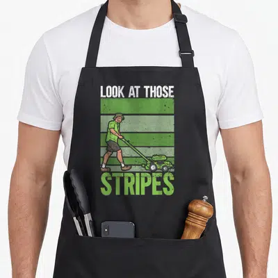 Those stripe apron - Distressed man pushing mower over green white brown stripes with text — shown on a black cotton canvas apron against a white background