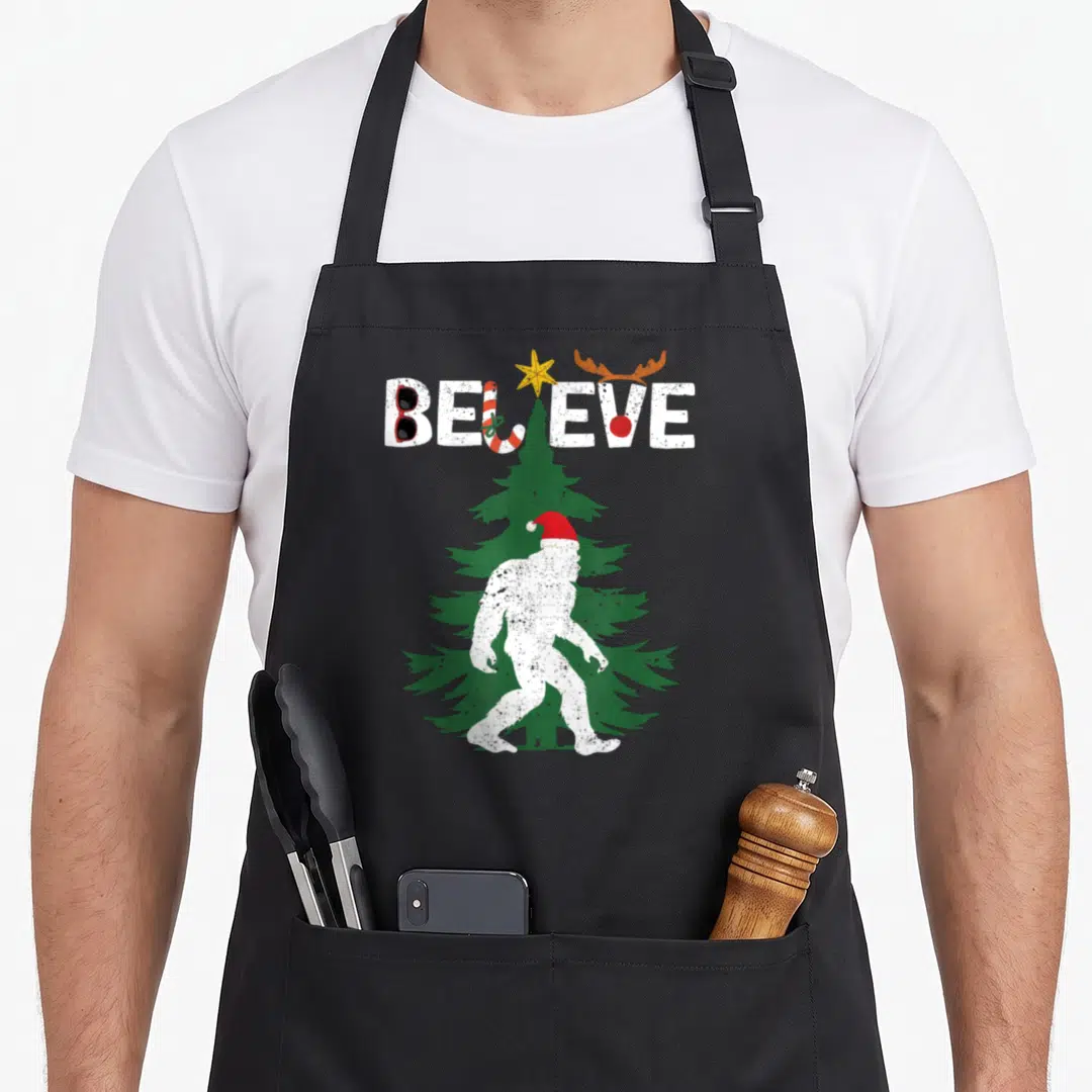 Santa yeti apron - Walking Bigfoot in Santa hat before green tree with distressed BELIEVE text and holiday icons — shown on a black cotton canvas apron against a white background