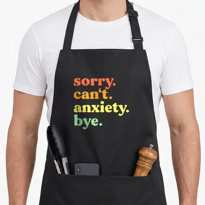 Mental anxious apron - Stacked typography reading sorry can't anxiety bye in orange yellow green — shown on a black cotton canvas apron against a white background