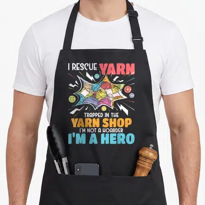 Rescue knitting apron - I Rescue Yarn text with colorful yarn balls and buttons in comic burst style — shown on a black cotton canvas apron against a white background