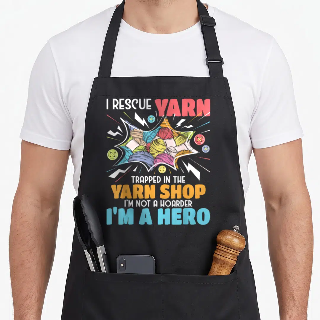 Rescue knitting apron - I Rescue Yarn text with colorful yarn balls and buttons in comic burst style — shown on a black cotton canvas apron against a white background