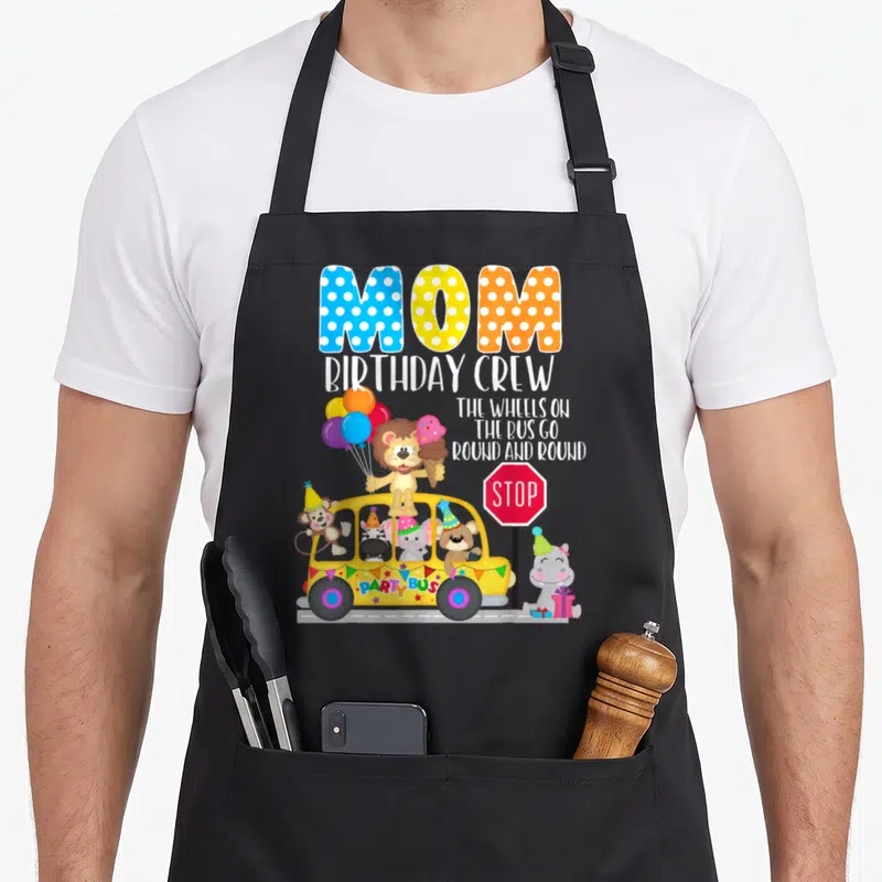 Wheel birthday apron - MOM BIRTHDAY CREW text above yellow party bus with lion hippo and balloons — shown on a black cotton canvas apron against a white background