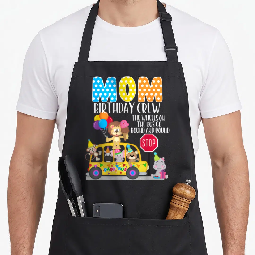 Wheel birthday apron - MOM BIRTHDAY CREW text above yellow party bus with lion hippo and balloons — shown on a black cotton canvas apron against a white background