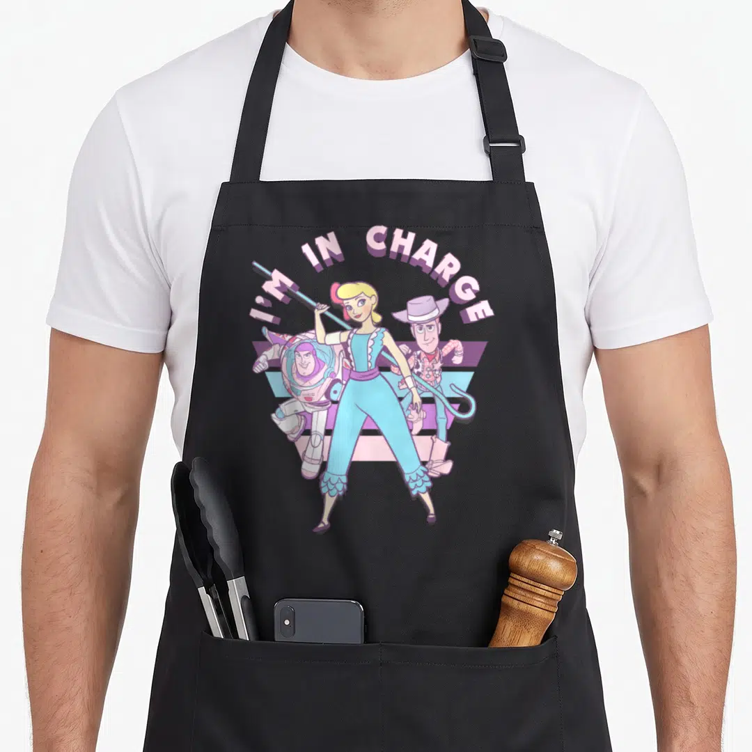 Charge little bo peep apron - I'M IN CHARGE text arching over Jessie, Buzz, and Woody with striped purple and blue — shown on a black cotton canvas apron against a white background