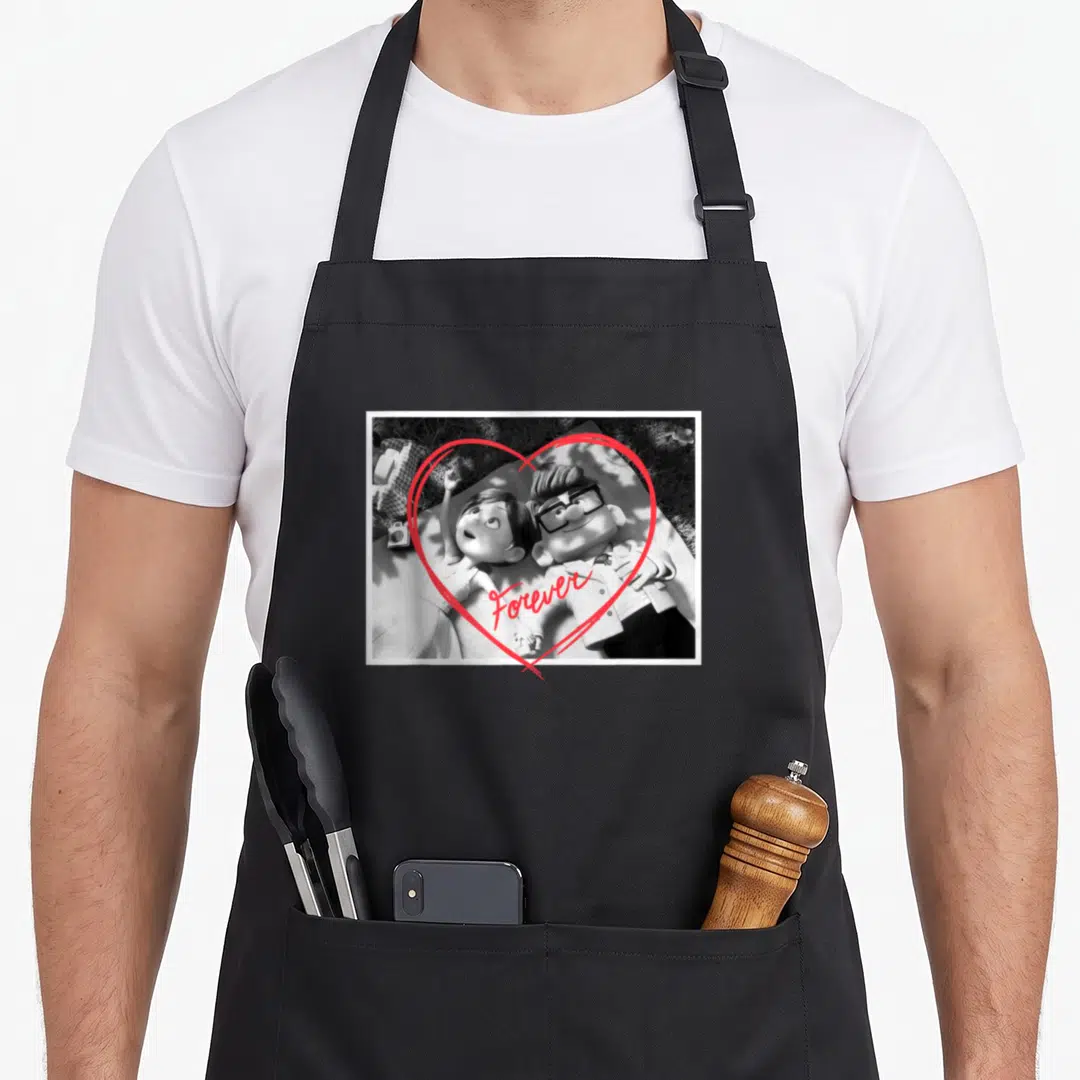 Forever carl and ellie apron - Black and white photo of couple lying on grass with red heart outline and Forever text — shown on a black cotton canvas apron against a white background