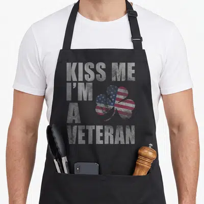 Kiss st patrick day aprons - Distressed KISS ME I'M A VETERAN text beside a shamrock filled with US flag pattern — shown on a black cotton canvas apron against a white background