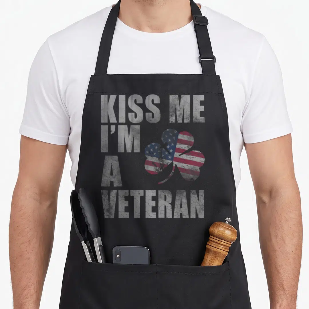 Kiss st patrick day aprons - Distressed KISS ME I'M A VETERAN text beside a shamrock filled with US flag pattern — shown on a black cotton canvas apron against a white background