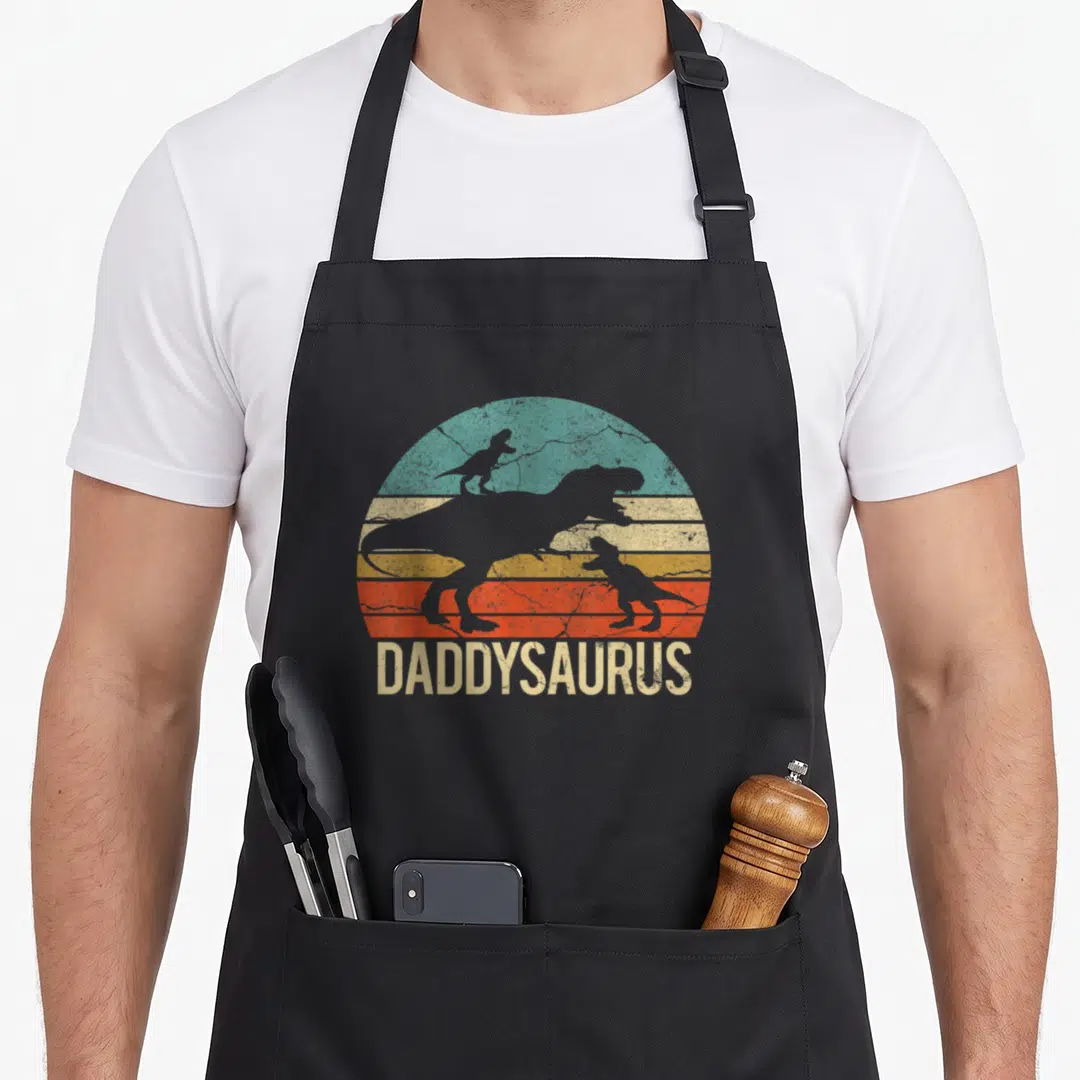Dinosaur daddy apron - Retro Daddysaurus text below three dinosaur silhouettes over a striped sun in teal, yellow — shown on a black cotton canvas apron against a white background