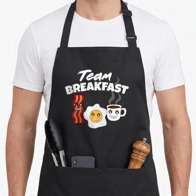 Breakfast egg apron - Cartoon bacon, fried egg, and coffee cup with smiling faces under Team BREAKFAST text — shown on a black cotton canvas apron against a white background