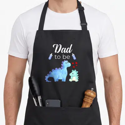 Baby dinosaur apron - Dad to be text with blue and teal watercolor dinosaurs and red hearts — shown on a black cotton canvas apron against a white background