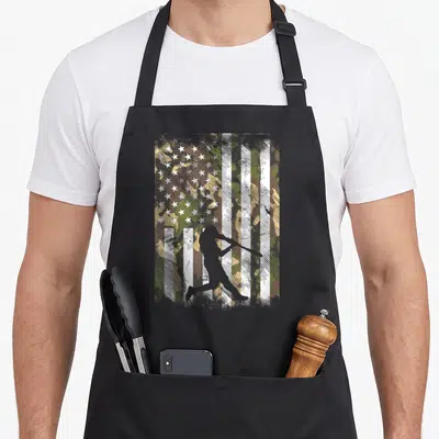 Baseball camo apron - White batter silhouette over distressed green and brown camouflage US flag pattern — shown on a black cotton canvas apron against a white background