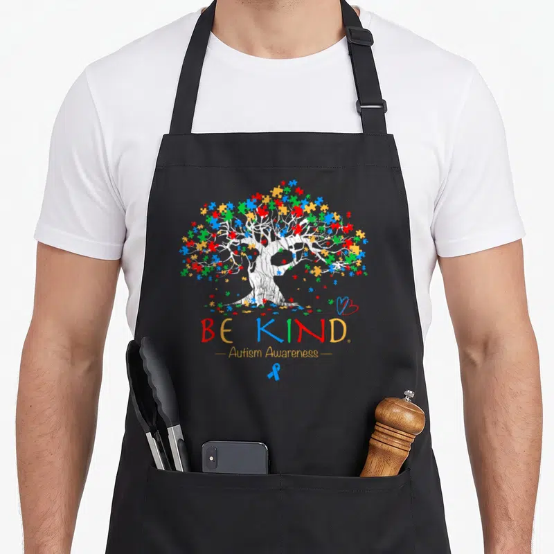 Awareness tree apron - White tree with red blue yellow green puzzle leaves and Be Kind text below — shown on a black cotton canvas apron against a white background