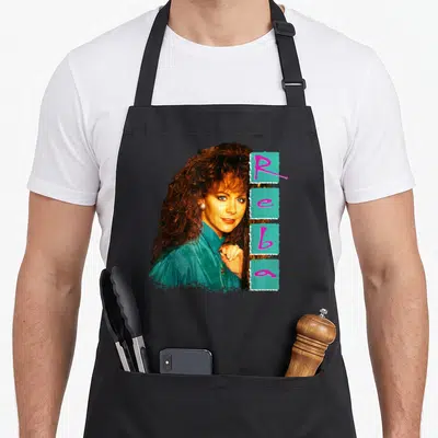 Reba cook's country aprons - Vintage photo of a woman next to vertical Reba text on teal blocks — shown on a black cotton canvas apron against a white background