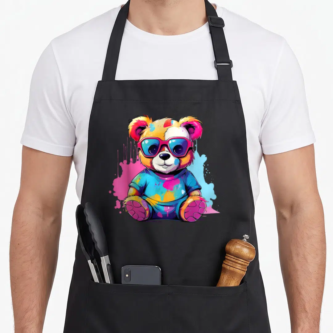Funny colorful teddy bear apron - Cartoon bear wearing sunglasses surrounded by blue, pink, yellow, and orange paint — shown on a black cotton canvas apron against a white background