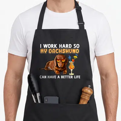 Cute dachshund apron - Brown dachshund with orange sunglasses lying next to cocktail glass with text above and below — shown on a black cotton canvas apron against a white background