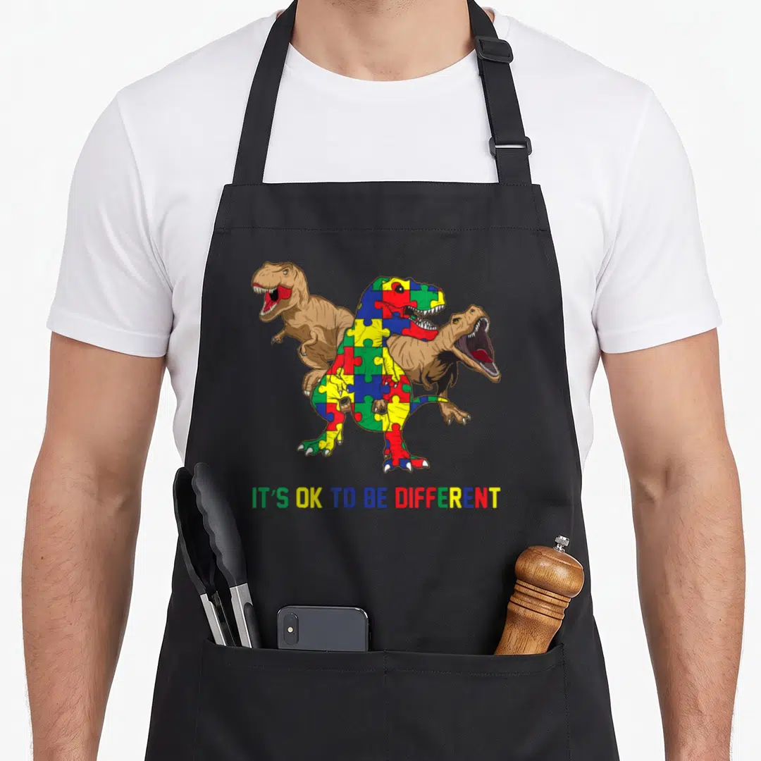 Autism awareness dinosaur apron - Three cartoon T-Rexes with central puzzle piece dinosaur and rainbow text IT'S OK — shown on a black cotton canvas apron against a white background