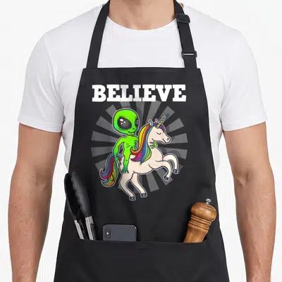 Alien unicorn apron - BELIEVE text above green alien riding white unicorn with rainbow mane on grey sunburst — shown on a black cotton canvas apron against a white background