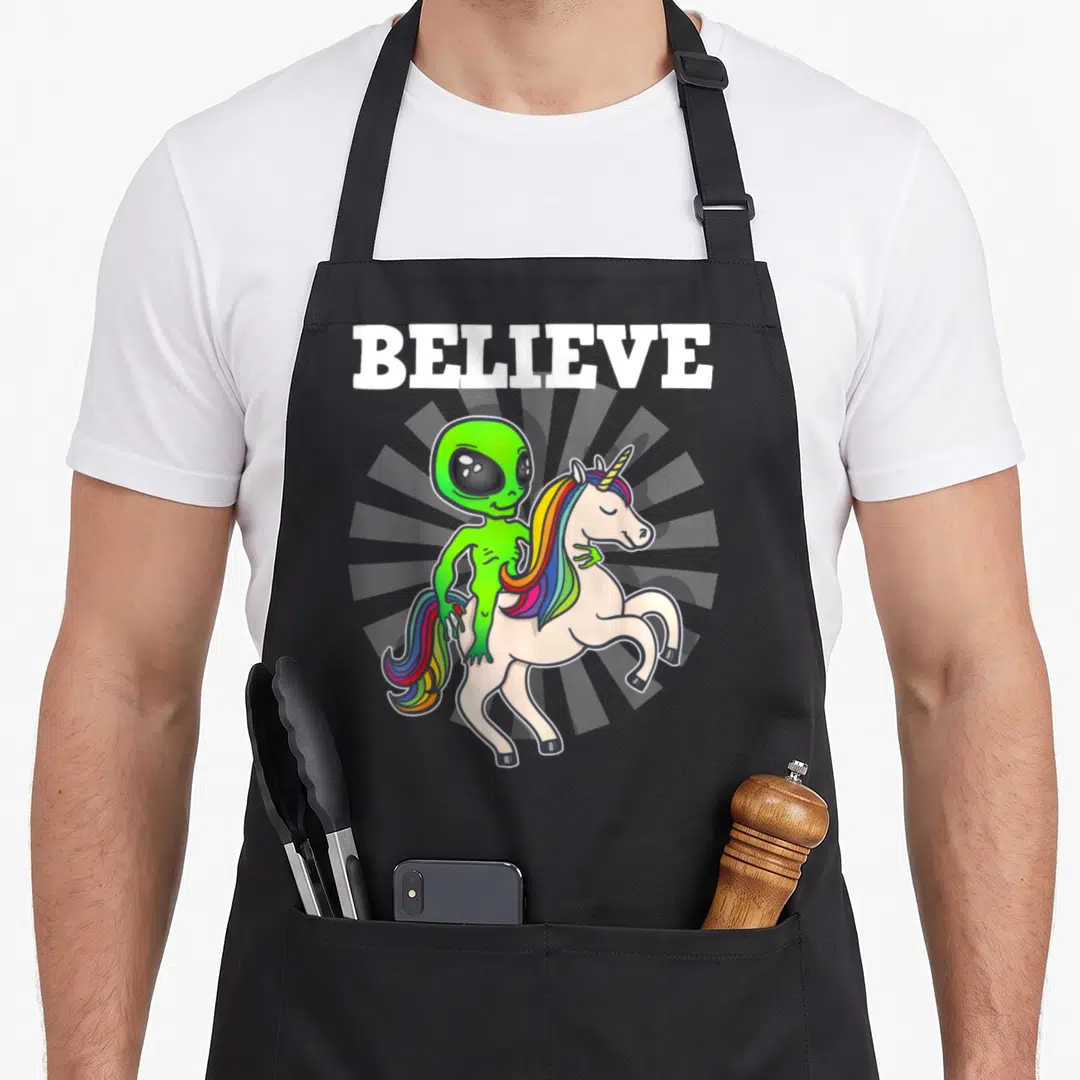Alien unicorn apron - BELIEVE text above green alien riding white unicorn with rainbow mane on grey sunburst — shown on a black cotton canvas apron against a white background