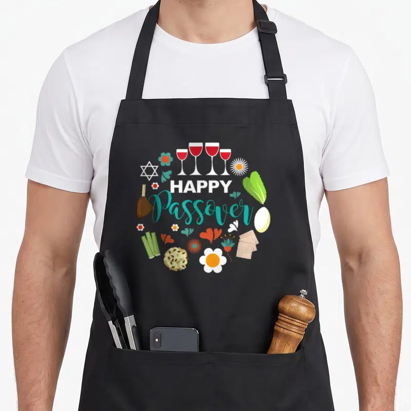Meal passover apron - HAPPY Passover text surrounded by circular Seder symbols and flowers in teal, red, and green — shown on a black cotton canvas apron against a white background