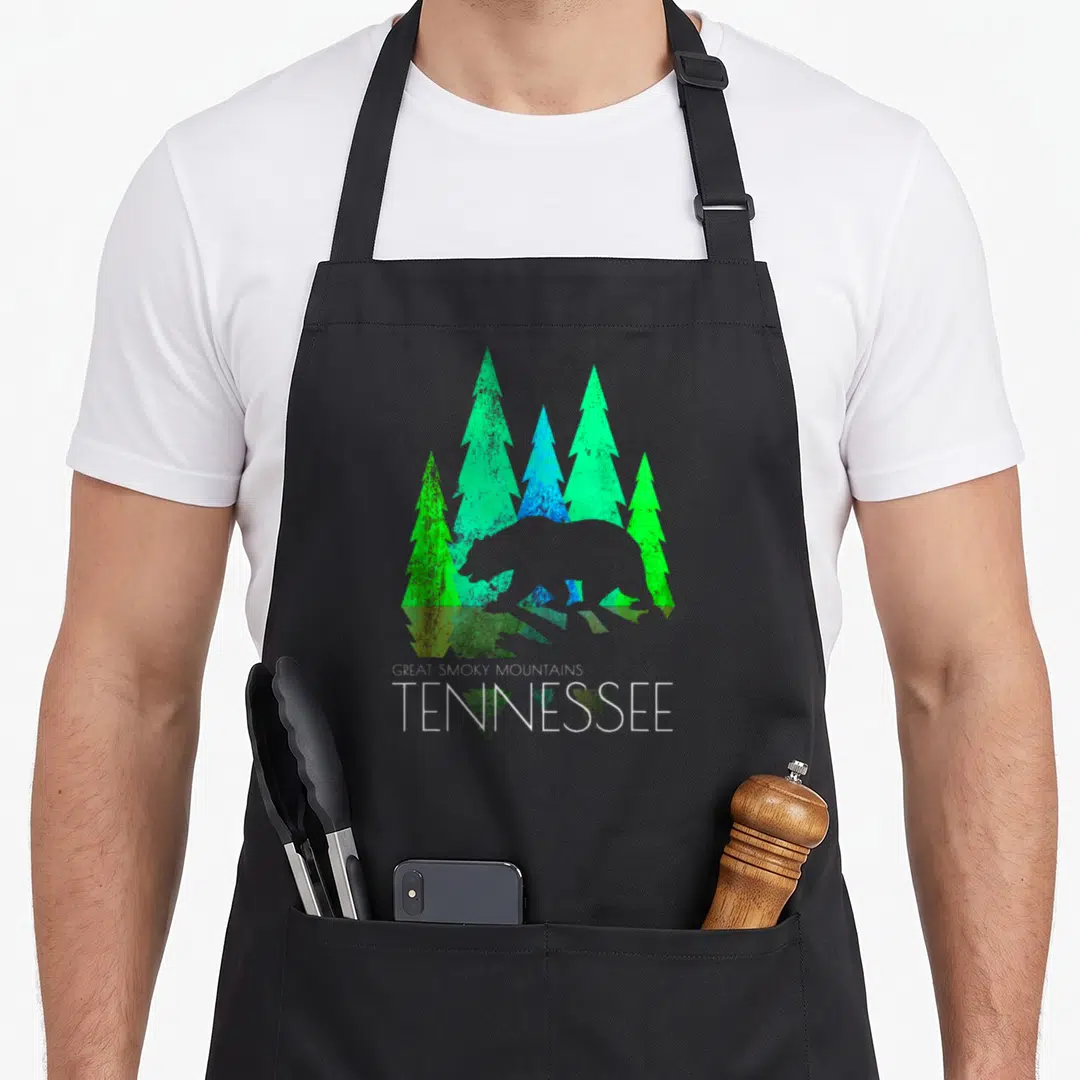Tennessee bear apron - Minimalist bear silhouette behind green and blue pine trees with stacked Great Smoky Mountains — shown on a black cotton canvas apron against a white background