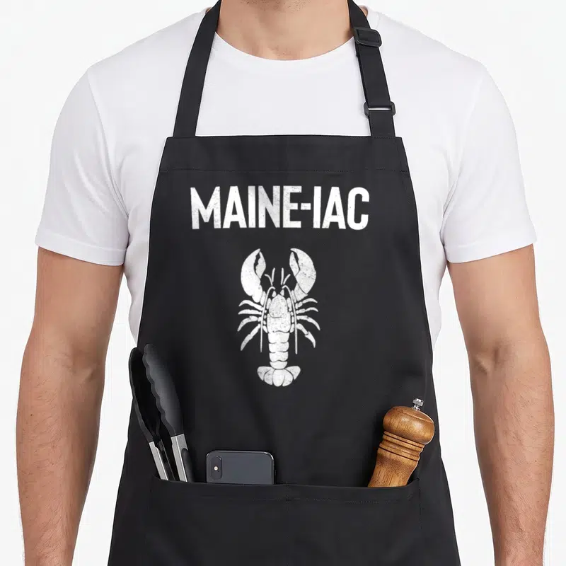 Maine fishing aprons - White distressed MAINE-IAC text above a white lobster illustration on a dark background — shown on a black cotton canvas apron against a white background