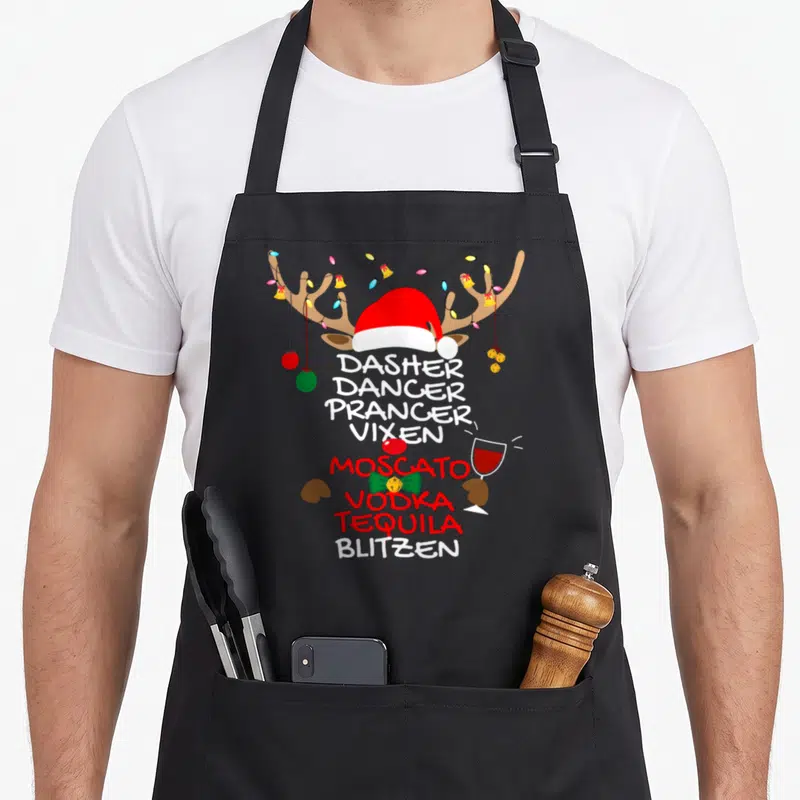 Vixen moscato apron - Cartoon reindeer head with Santa hat, antler lights, and stacked text listing Dasher Dancer — shown on a black cotton canvas apron against a white background