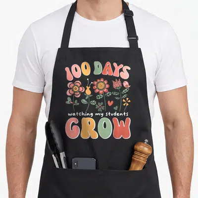 Growing boho apron - Arched 100 DAYS text above cartoon flowers, bird, hearts, and stacked GROW lettering in pink — shown on a black cotton canvas apron against a white background