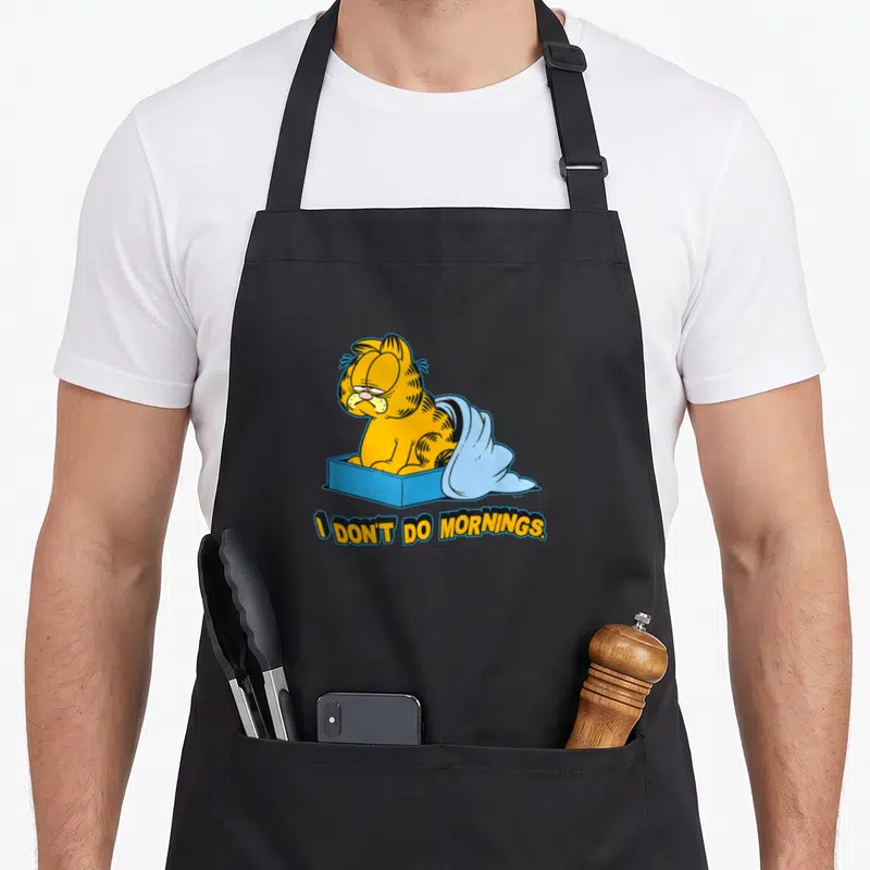Funny morning routine apron - Orange cartoon cat in blue box with light blue blanket and text I DON'T DO MORNINGS — shown on a black cotton canvas apron against a white background