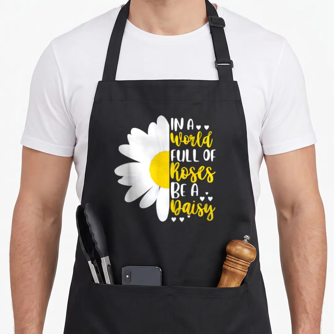 Rose daisy apron - Half white daisy with yellow center next to stacked white and yellow text with small hearts — shown on a black cotton canvas apron against a white background