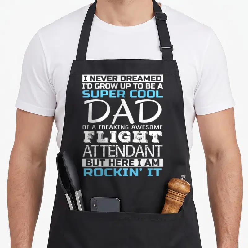 Funny flight attendant apron - White and blue stacked text reading super cool dad of a freaking awesome flight — shown on a black cotton canvas apron against a white background
