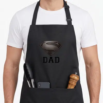 Steel dad mode apron - Realistic brown shield emblem above black DAD text — shown on a black cotton canvas apron against a white background