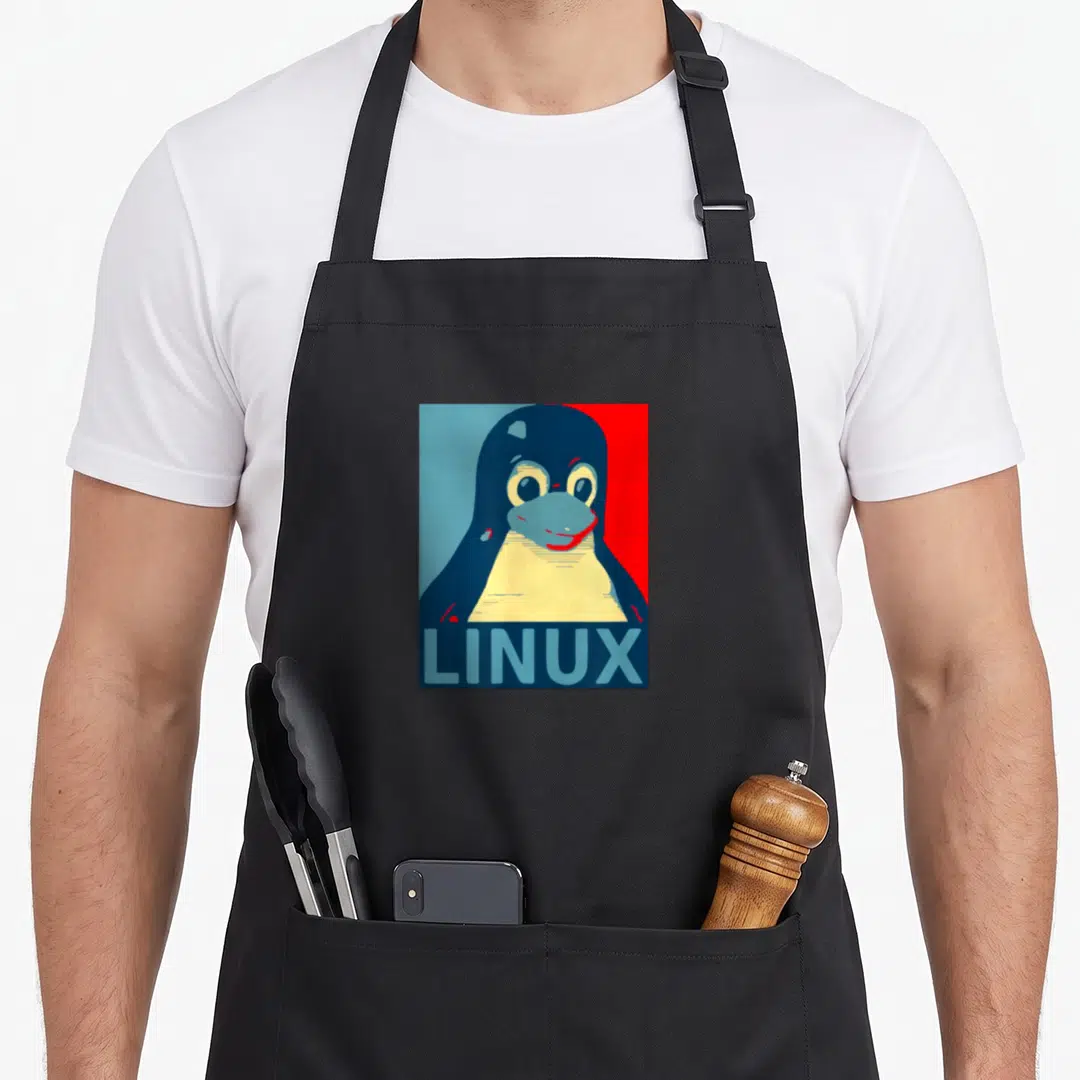 Linux tux penguin aprons - Cartoon penguin portrait above LINUX text on split blue and red background — shown on a black cotton canvas apron against a white background