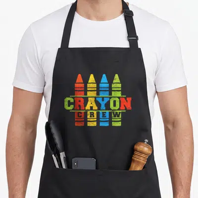 Cray crew apron - Four red yellow blue green crayons with CRAYON CREW text — shown on a black cotton canvas apron against a white background