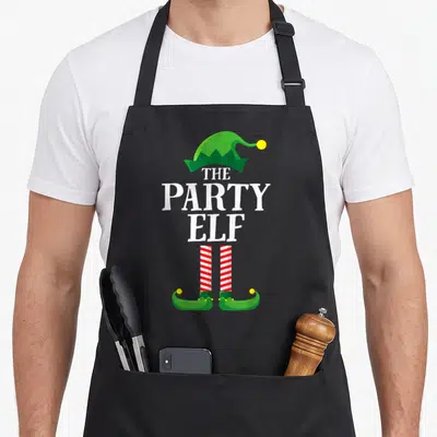 Vintage family christmas elf apron - Green elf hat, THE PARTY ELF text, red white striped legs, green shoes with yellow — shown on a black cotton canvas apron against a white background