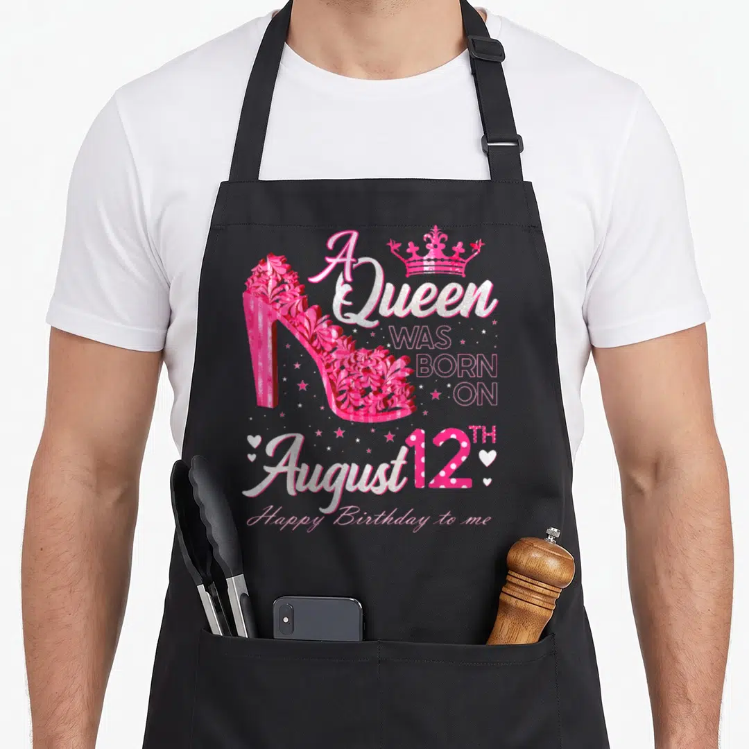 August heel birthday apron - Pink floral high heel with A Queen Was Born on August 12TH text and crown — shown on a black cotton canvas apron against a white background