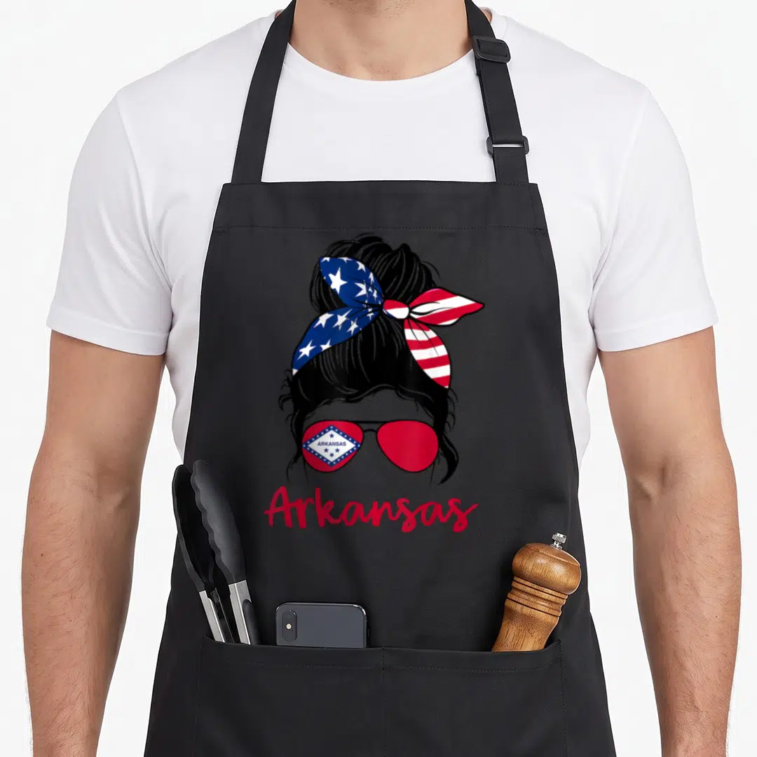 Bow messy aprons - Cartoon woman bun with USA flag bow and Arkansas state flag sunglasses — shown on a black cotton canvas apron against a white background