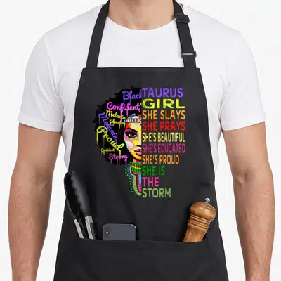 Taurus she birthday apron - Stylized woman’s face beside stacked text TAURUS GIRL SHE SLAYS SHE IS THE STORM in yellow — shown on a black cotton canvas apron against a white background