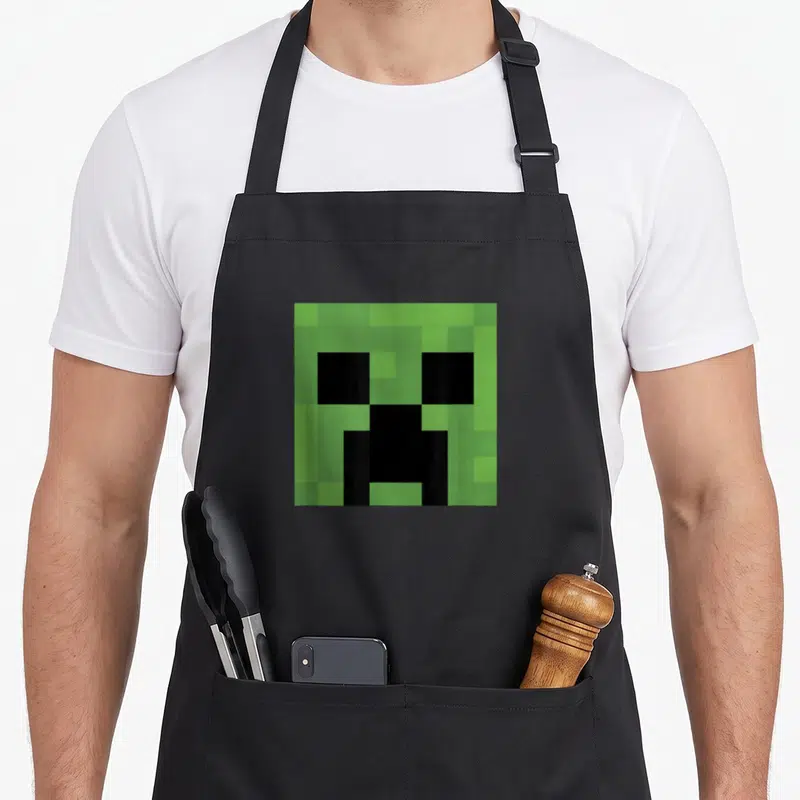 Creeper face apron - Green and black pixelated square monster face centered on a black bib apron — shown on a black cotton canvas apron against a white background
