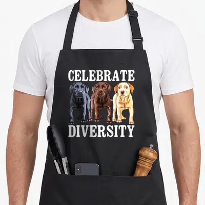 Celebrate labrador apron - Three cartoon dogs in black, brown, and yellow standing under celebrate diversity text — shown on a black cotton canvas apron against a white background