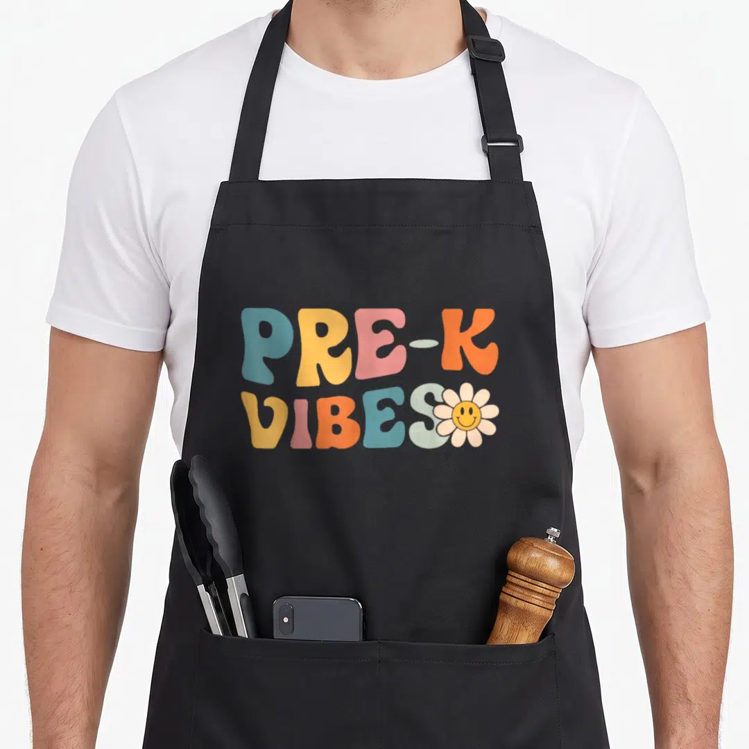 Kindergarten pre back apron - PRE-K VIBES retro text in blue yellow pink orange with a smiley face flower on the right — shown on a black cotton canvas apron against a white background