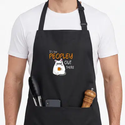 Peopley grumpy cat apron - Cartoon white cat holding orange mug surrounded by text It's too PEOPLEY OUT THERE — shown on a black cotton canvas apron against a white background