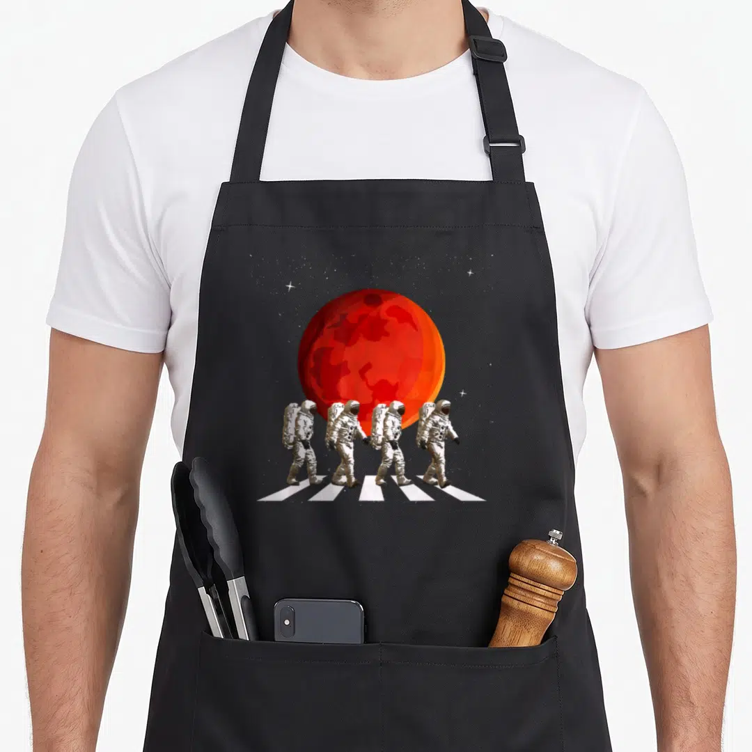 Spaceman walking apron - Four astronauts walking on white stripes in front of a red planet and stars — shown on a black cotton canvas apron against a white background